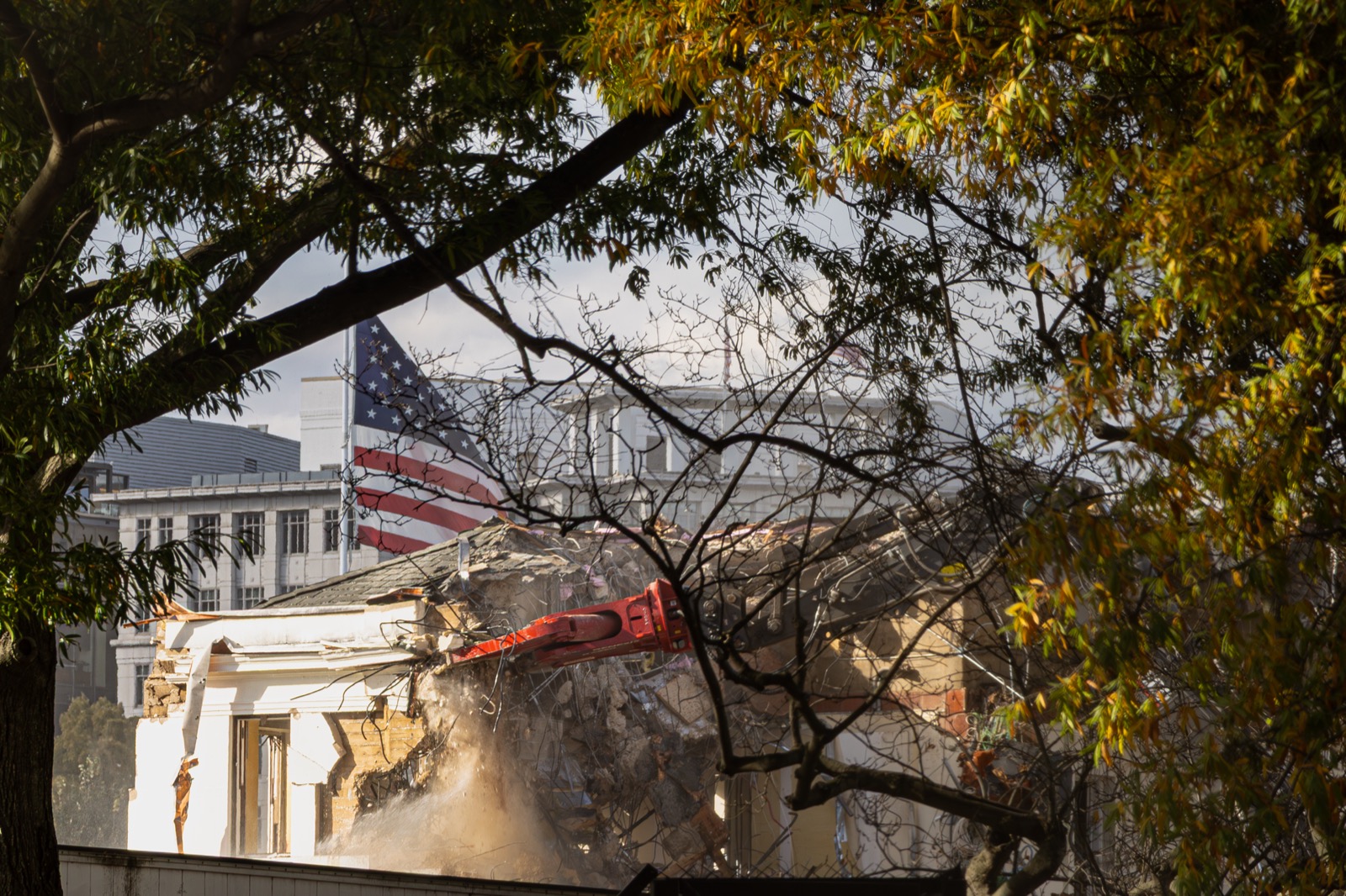 An excavator arm tears into the partially demolished White House East Wing, an American flag visible through the dust and autumn trees