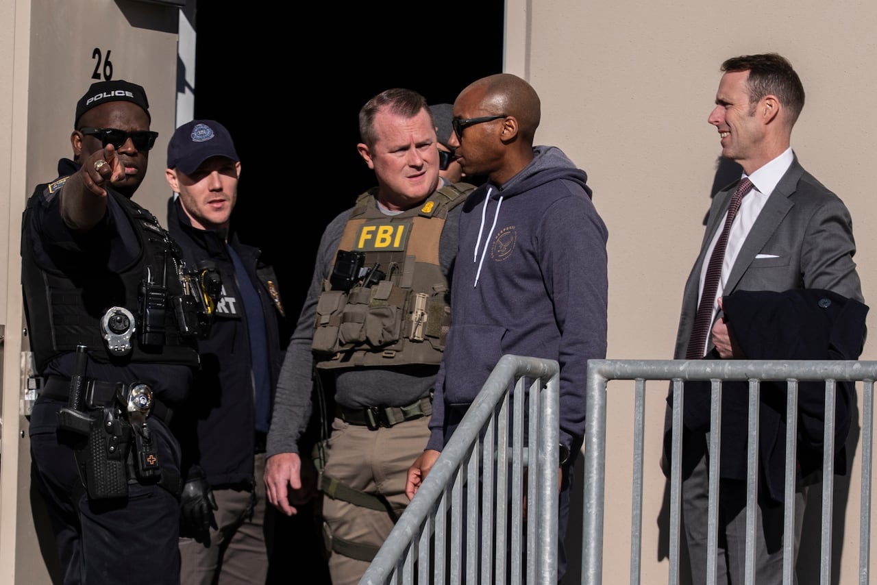 FBI agents in tactical vests and local police officers stand outside the Fulton County Election Hub on January 28, 2026. An agent wearing an FBI vest speaks with others near building entrance marked '26'.