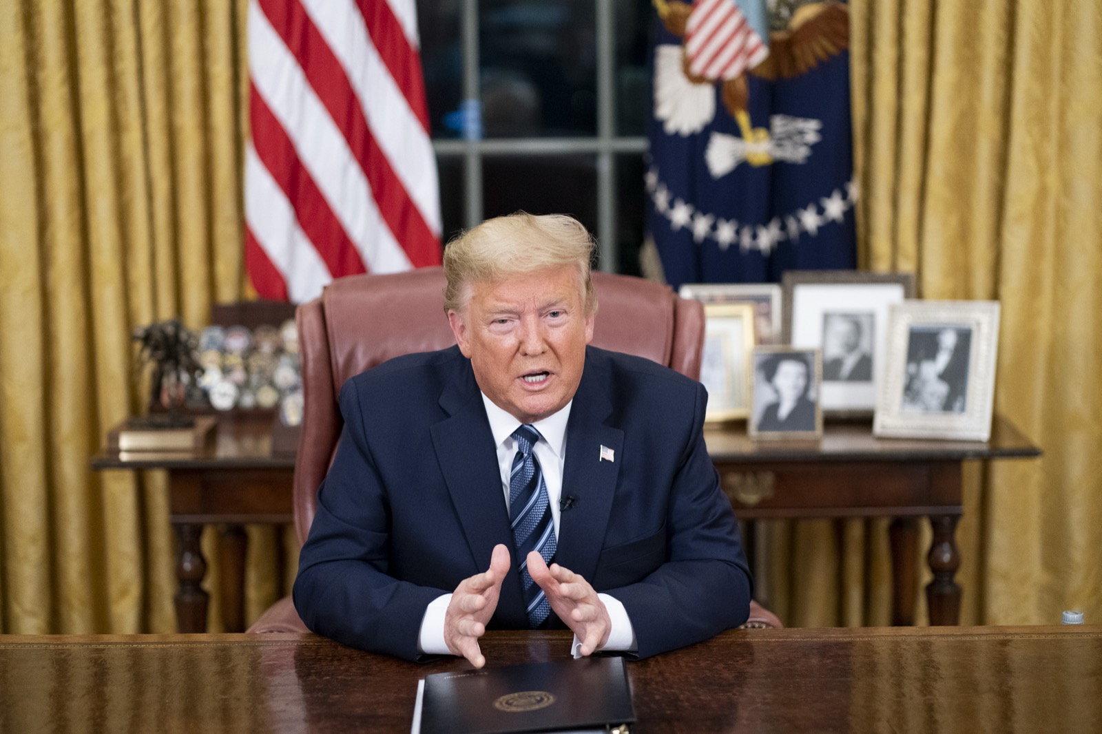 Donald Trump seated at the Resolute Desk in the Oval Office, mid-sentence, flags behind him, delivering a televised address