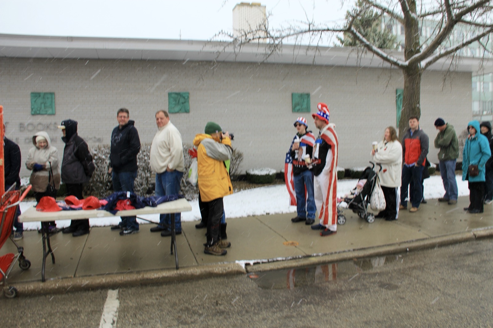 Supporters in winter coats wait in falling sleet outside a Trump rally in Racine, Wisconsin, one wearing an Uncle Sam costume, beside a folding table of red campaign hats