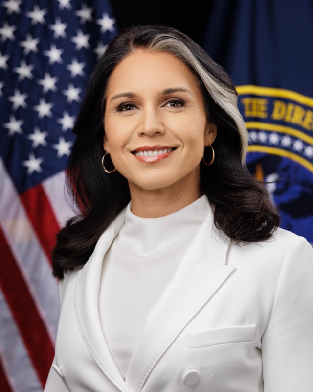 Official portrait of Tulsi Gabbard as Director of National Intelligence. She wears a white blazer and stands before an American flag and the DNI seal.